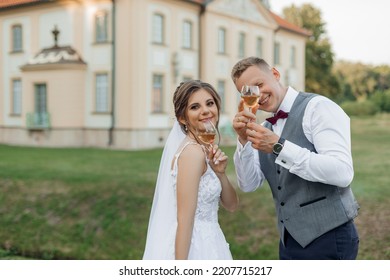 Portrait Of Amazing Wedding Couple Standing Near Old Building In Park In Summer. Young Man Groom With Young Attractive Woman Bride Raising Hands With Glasses Of Wine. Relationship, Love, Celebration.