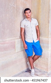 Portrait Of American College Student, Wearing Iron Short Sleeve Shirt, Blue Cargo Shorts, Sneakers, Standing Against Wall In Corner On Campus In Hot Summer, Relaxing. Filtered Look With Purple Tint.