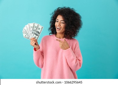 Portrait of american successful woman 20s with afro hairstyle holding lots of money dollar ï»¿banknotes isolated over blue background