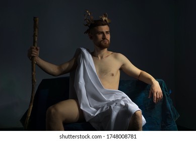 Portrait of the ancient Greek god Olympus, in white clothes, with a wreath on his head, holds a staff, sitting on a dark bench. On a gray background.