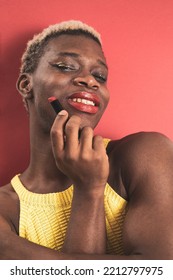 A Portrait Of An Androgynous Black Man On A Red Background While Putting On Lipstick.