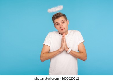 Portrait Of Angelic Nice-looking Man With Halo Above Head Holding Hands In Prayer And Looking At Camera With Timid Shy Expression, Obedient Behavior. Indoor Studio Shot Isolated On Blue Background