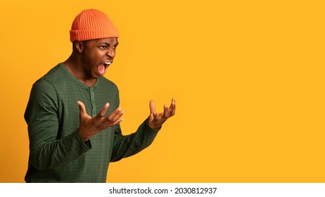 Portrait Of Angry African American Man Screaming With Rage, Furious Agressive Young Black Guy Raising Hands And Shouting While Standing Over Yellow Background In Studio, Panorama With Copy Space