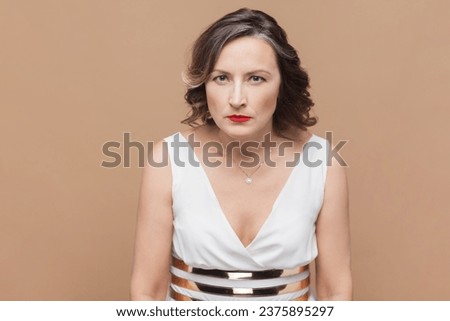 Portrait of angry aggressive middle aged woman with wavy hair looking attentively at camera with hate, wearing white dress. Indoor studio shot isolated on light brown background.