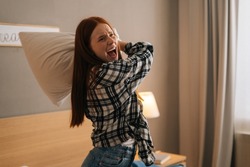 Portrait Of Angry Annoyed Woman With Open Mouth Shouting And Yelling With Aggressive Expression, Throwing Pillow Sitting On Bed In Bedroom. Furious Crazy Redhead Female Vented Anger In Privacy Of Home