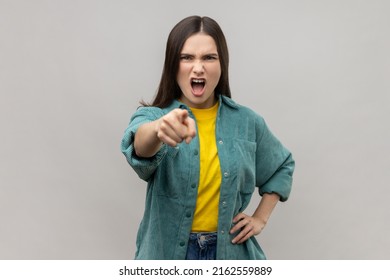 Portrait of angry bossy woman with dark hair pointing to camera with aggressive expression, making choice, wearing casual style jacket. Indoor studio shot isolated on gray background.