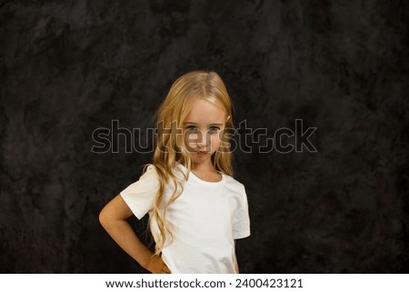 Portrait of angry cover girl kid model in white t-shirt expression emotion, frowning looking at camera, studio shot. Scowl child 6 year old posing at black. Kids emotional concept. Copy ad text space
