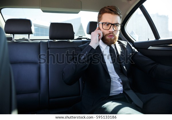 Portrait of\
an angry frustrated business man in eyeglasses talking on mobile\
phone while sitting in the back seat of\
car