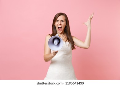 Portrait of angry irritated bride woman in wedding dress screaming in megaphone and spreading hands isolated on pink pastel background. Organization of wedding concept. Copy space for advertisement
