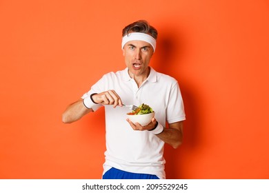 Portrait Of Angry Middle-aged Male Athlete, Eating Salad And Looking Disturbed, Standing Over Orange Background