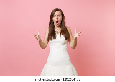 Portrait of angry shocked bride woman with opened mouth in white wedding dress standing spreading hands isolated on pink pastel background. Wedding celebration concept. Copy space for advertisement