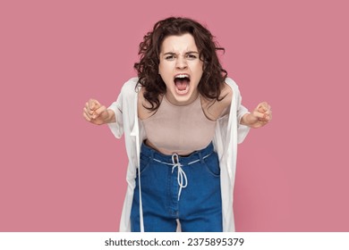 Portrait of angry woman with curly hair wearing casual style outfit standing arguing with somebody, clenched fists, screaming with hate and aggression. Indoor studio shot isolated on pink background.