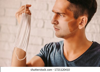 Portrait Of Angry Young Man With Funny Sunburn Lines Holding Medical Face Mask In Hand. Confused Hipster Man With Sun Tanned Lines On Face After Wearing Protective Face Mask. COVID-19 Prevention