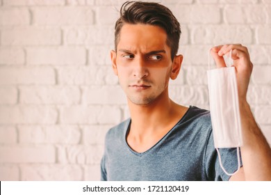 Portrait Angry Young Man With Funny Sun Tanned Lines On Face After Wearing Medical Face Mask. Confused Hipster Man With Sunburn Tan Face Skin Holding Protective Face Mask In Hand. COVID-19 Quarantine