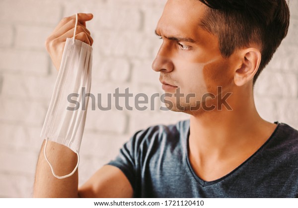 Portrait of angry young man with funny sunburn\
lines holding medical face mask in hand. Confused hipster man with\
sun tanned lines on face after wearing protective face mask.\
COVID-19 prevention
