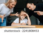 Portrait of angry young parents yelling and scolding together lazy little daughter sitting at table, doing homework, sad looking at camera. Concept of parent disciplining child for bad education.