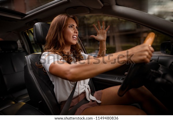 Portrait of angry young\
sitting woman pissed off by drivers in front of her and gesturing\
with hands. 