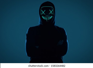 Portrait Of An Anonymous Man In A Black Hoodie Hiding His Face Behind A Scary Neon Mask. Studio Shot.