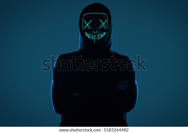 怖いネオンマスクの後ろに顔を隠す黒いフードを着た匿名の男性のポートレート スタジオショット の写真素材 今すぐ編集