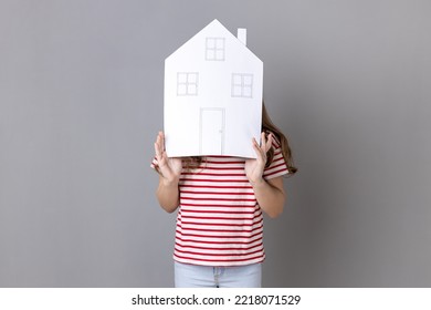 Portrait Of Anonymous Unknown Little Girl Wearing Striped T-shirt Covering His Face With Big Paper House, Advertising Accommodation. Indoor Studio Shot Isolated On Gray Background.