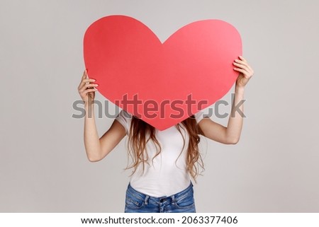 Portrait of anonymous woman hiding her face behind big read paper heart, holding symbol of love, care, generosity, give hope, wearing white T-shirt. Indoor studio shot isolated on gray background.