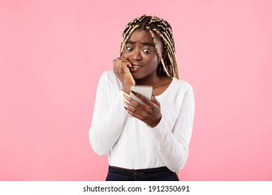 Portrait Of Anxious Scared Black Young Woman Looking At Mobile Phone Seeing Bad News, Photos Or Message. Worried Lady Biting Nails Isolated On Pink Studio Wall Background. Human Reaction, Expression