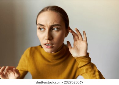 Portrait of anxious scared female stepping carefully eavesdropping with anxious facial expression, listening to strange scary sounds attentively, holding hand around ear isolated on gray background