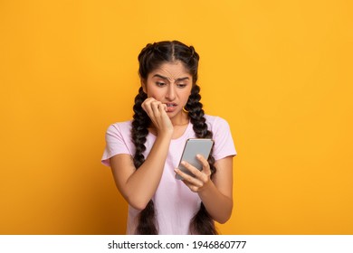 Portrait Of Anxious Scared Indian Young Woman Looking At Mobile Phone Seeing Bad News, Photos Or Message. Worried Lady Biting Nails Isolated On Yellow Studio Background. Human Reaction, Expression