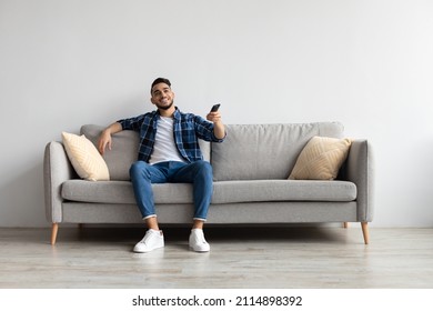 Portrait of Arab guy watching TV show or film, holding remote control, switching channels. Young smiling man spending weekend free time sitting on comfortable sofa at home in living room, copy space