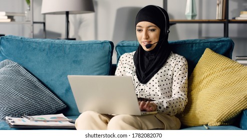 Portrait Of Arabic Business Woman At Home Working Remotely While Sitting On Sofa In Living Room With Headset And Laptop Computer.