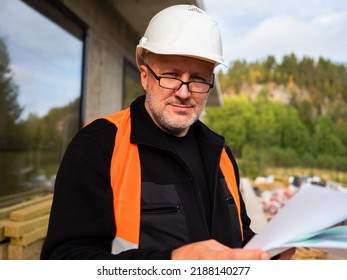 Portrait Architect. Architect With Papers Stands Outdoor. Man In Construction Helmet. Guy In Orange Vest Is Looking At Camera. Employee Architectural Company Inspects Construction. 