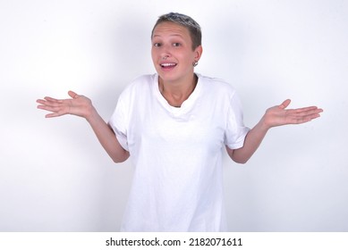 So what? Portrait of arrogant young woman with short hair wearing white t-shirt over white background shrugging hands sideways smiling gasping indifferent, telling something obvious.