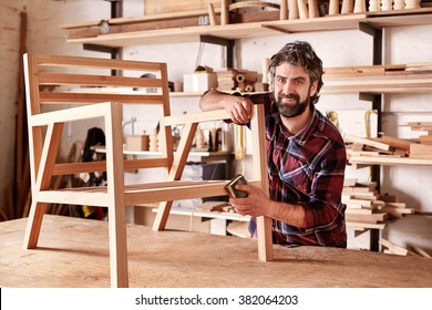 Portrait Of An Artisan Designer, With New Piece Of Furniture, Finishing Off The Sanding Of The Chair In His Studio, With Shelves Of Wood Behind Him