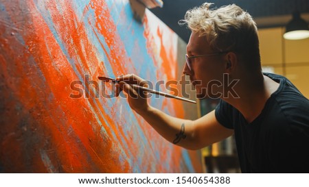 Portrait of Artist Working on Abstract Painting, Uses Paint Brush To Create Daringly Emotional Modern Picture. Dark Creative Studio Large Canvas Stands on Easel Illuminated. Side View Close-up Shot 