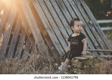 An Portrait Of Asian 1 Year Old Girl Is Chilling And Looking Over Her Shoulder Wearing Black Outfit And Sneakers In The Middle Of A Pile Of Old Wooden Pallet.