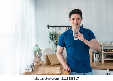 Portrait of Asian attractive male holding a glass of water in kitchen. Young thirsty handsome man pouring and drink clean mineral natural in cup after wake up in early morning for health care in house