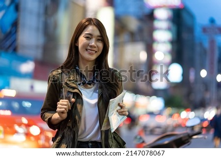 Portrait of Asian attractive woman backpacker travel in city at night. Young woman tourist traveler stand in beautiful street enjoy spend time on holiday vacation trip in Thailand and look at camera.