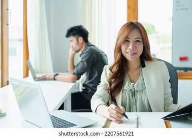 Portrait Of An Asian Bank Employee Using Notebooks To Take Notes, Budget Documents And Computers To Close Financial Statements.