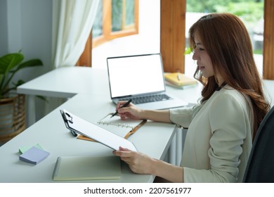 Portrait Of An Asian Bank Employee Using Notebooks To Take Notes, Budget Documents And Computers To Close Financial Statements.