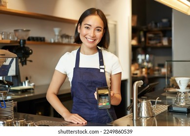 Portrait of asian barista girl at counter, showing card machine to client who wants to pay contactless, taking order, standing in cafe. - Powered by Shutterstock