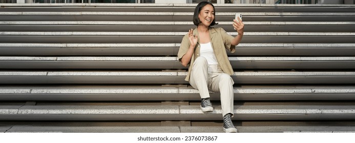 Portrait of asian beautiful girl takes selfie on smartphone. Young korean woman sits on stairs outdoors and makes photos on mobile phone. - Powered by Shutterstock