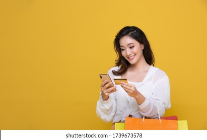 Portrait Asian Beautiful Happy Young Woman Smiling Cheerful And She Is Holding Credit Card And Using Smartphone For Shopping Online With Shopping Bags Isolated On Yellow Studio Background.