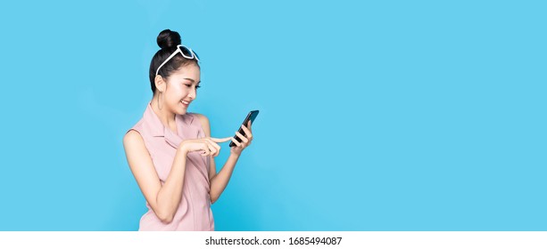 Portrait Of Asian Beautiful Young Woman Holding Smartphone And Pointing Finger On Mobile Phone With Smile Face. The Girl Standing And Looking At Phone In Studio. Advertisement & Presentation Concept.