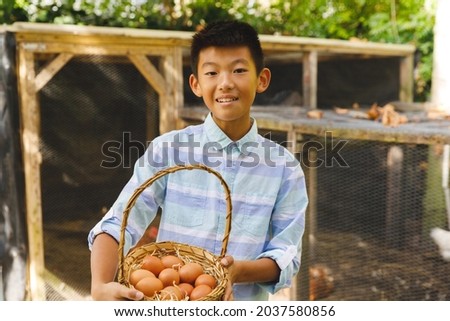 Portrait of asian boy smiling and holding basket, collecting eggs from hen house in garden. organic produce and self sufficiency at home.