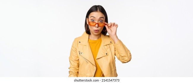 Portrait of asian brunette woman in stylish sunglasses, looks surprised and impressed at camera, checking out big news, wow face expression, standing over white background
