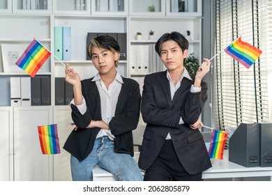 Portrait Of Asian Businessman Gay Working In Office, Holding LGBT Flag. Attractive Male Lgbtq Collegues In Formal Wear Sitting On Table, And Look At Camera With Gay Pride And Rainbow Flag At Workplace