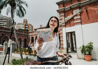 Portrait of asian chinese woman tourist hipster standing with map and bicycle in taichung city center in taiwan. Traveling summer vacation concept. young girl traveler in glasses reading paper guide - Powered by Shutterstock