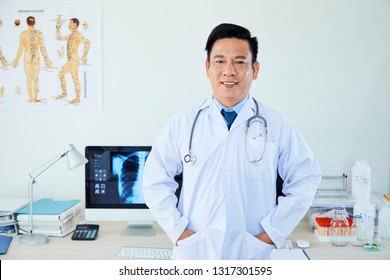 Portrait Of Asian Confident Mature Neurologist Standing In Lab Coat At His Office And Smiling At Camera