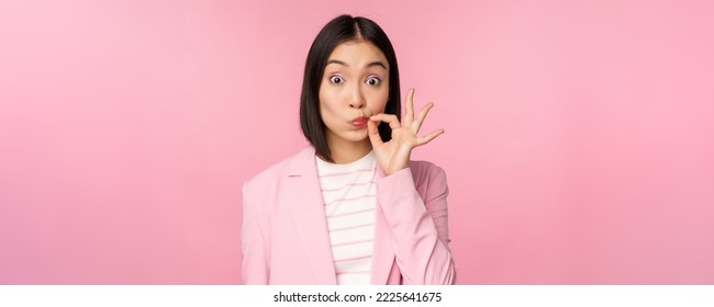 Portrait of asian corporate woman showing mouth seal, close shut lips on key gesture, promise keep secret, standing over pink background in suit. - Shutterstock ID 2225641675