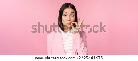 Portrait of asian corporate woman showing mouth seal, close shut lips on key gesture, promise keep secret, standing over pink background in suit.